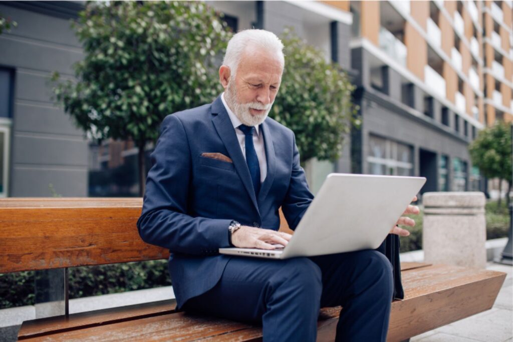 Older man in business attire working on laptop on bench outdoors